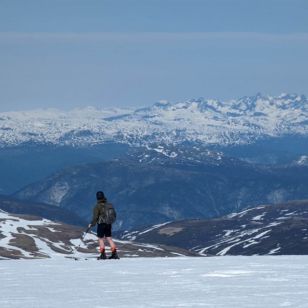 ein mann står på ein fjelltopp i shorts, og med ski på beina, og ser på utsikta. I bakgrunnen ser ein nokre spisse fjell som er langt, langt, unna.
