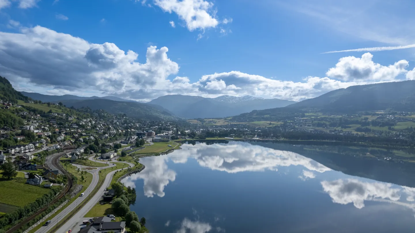 Dronefoto av Vangsvatnet og Voss sentrum en sommersdag. Blå himmel og noen skyer speiler seg i vannet. 